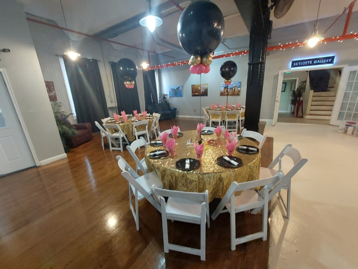 Round banquet table with gold sequin linen, pink napkins, and black-and-gold balloon centerpieces inside SkyLofts Gallery party venue set for a celebration event.