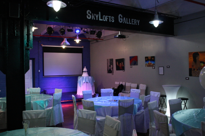 Reception setup at SkyLofts Gallery featuring teal-draped tables, white chair covers, and a cake table beneath a projector screen—perfect for weddings and other events.